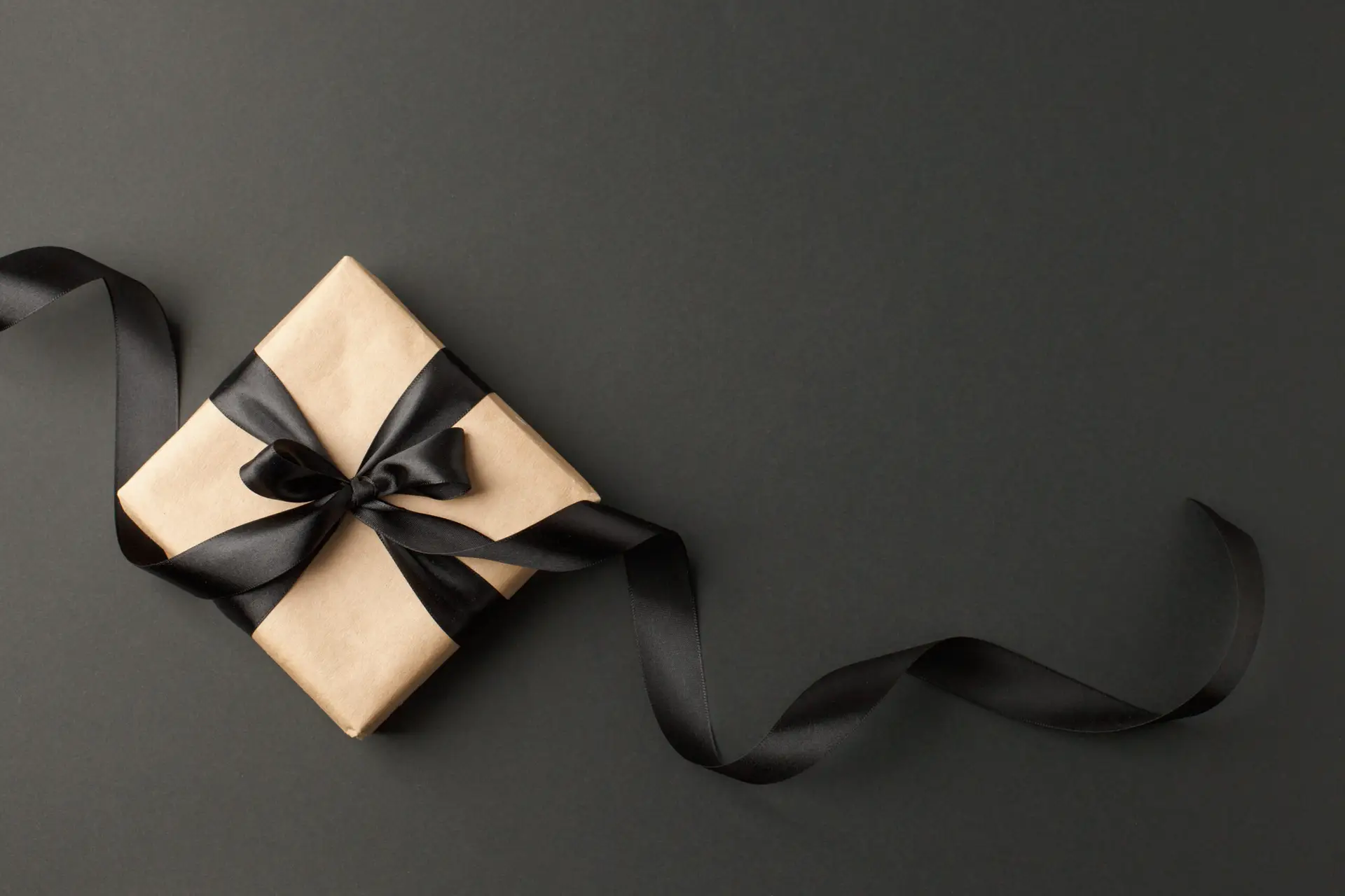 photo of tan gift box wrapped with a long black ribbon on black background